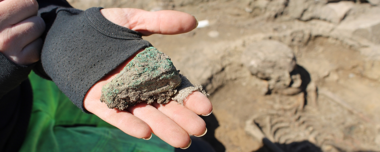 A person holds a triangle-shaped bronze ornament with the excavation of a skeleton in the background.