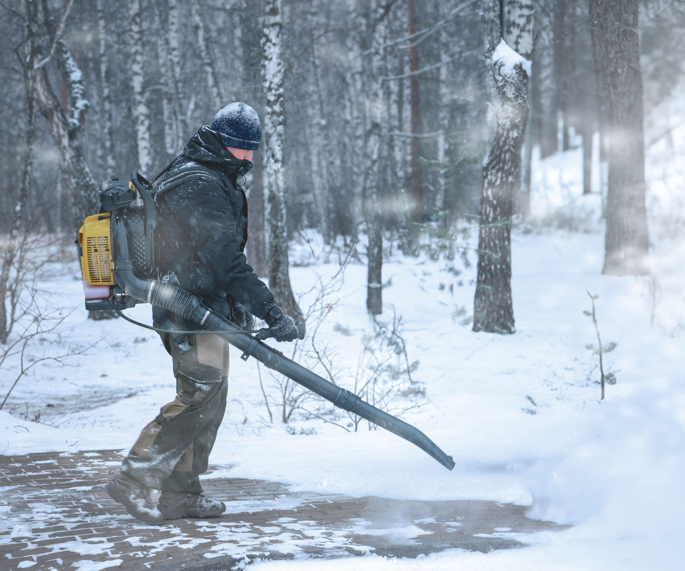 A worker is using a leaf blower to clear snow in a forest, wearing thick winter clothing