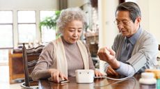 An elderly woman helps an elderly man check his blood pressure.