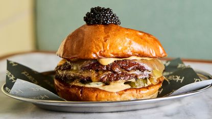 A double smash burger bun with cheese and pickle filling and caviar placed on top sits in a chrome platter on branded oven paper in a green and butter yellow restaurant.