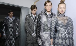 One female and three male models wearing looks from Moncler Gamme Bleu's collection. The male models are wearing shirts, ties, jackets, gloves and a one piece in varying shades of grey and white with a repeating diamond pattern and quilting. The female model is wearing a top and skirt with the same colours and pattern with a matching scarf around her neck