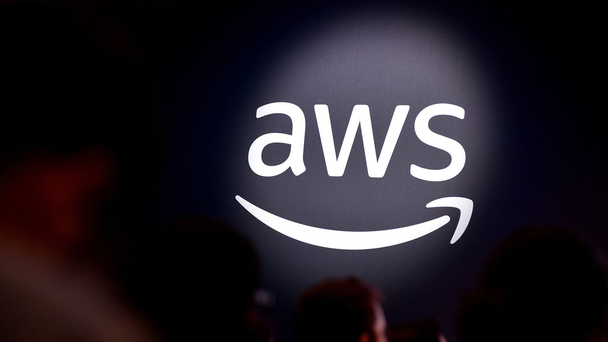 A photo of the AWS logo (the letters &quot;AWS&quot; with a curved arrow beneath moving from left to right) in white on a black wall, and lit by a spotlight. Closer to the camera and out of focus, a conference crowd is sat.