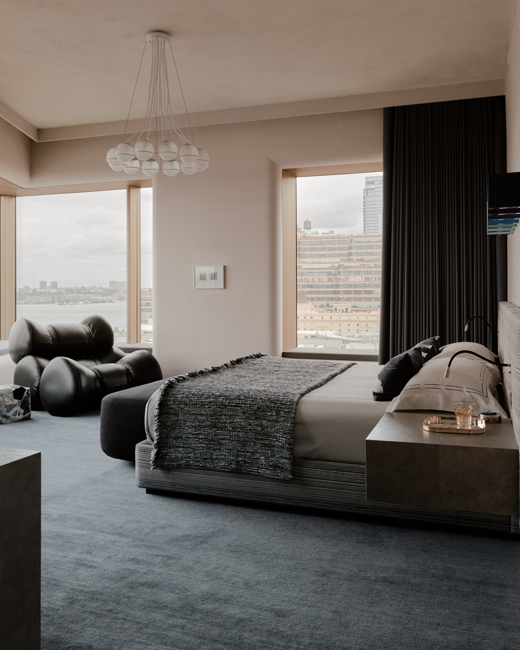 modern bedroom with dark carpet, gray bed, pendant light, beige walls, dark curtains, and a designer armchair in the corner
