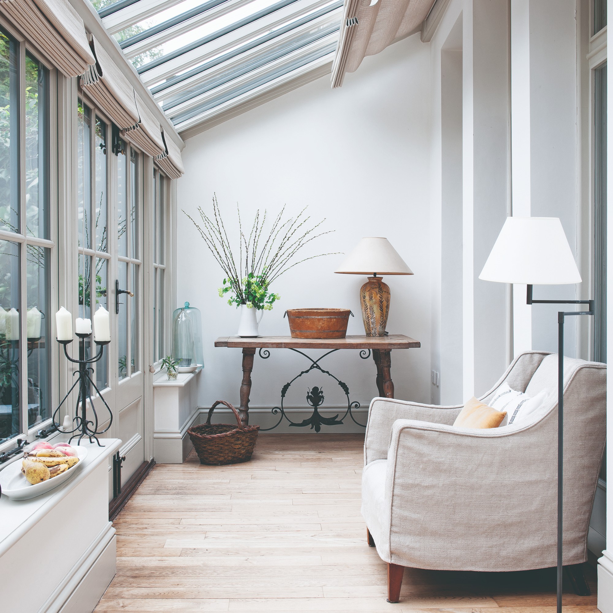 A small conservatory with Roman blinds covering the windows and a loose-cover cream-coloured armchair