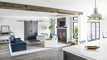 A view of a living room from the connecting kitchen, with large black barn doors, a blue velvet sofa and brick fireplace