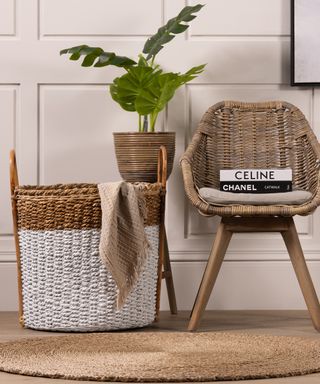 Large storage basket with blanket hanging over the edge in front of plant on stand and next to side chair with books from Where Saints Go
