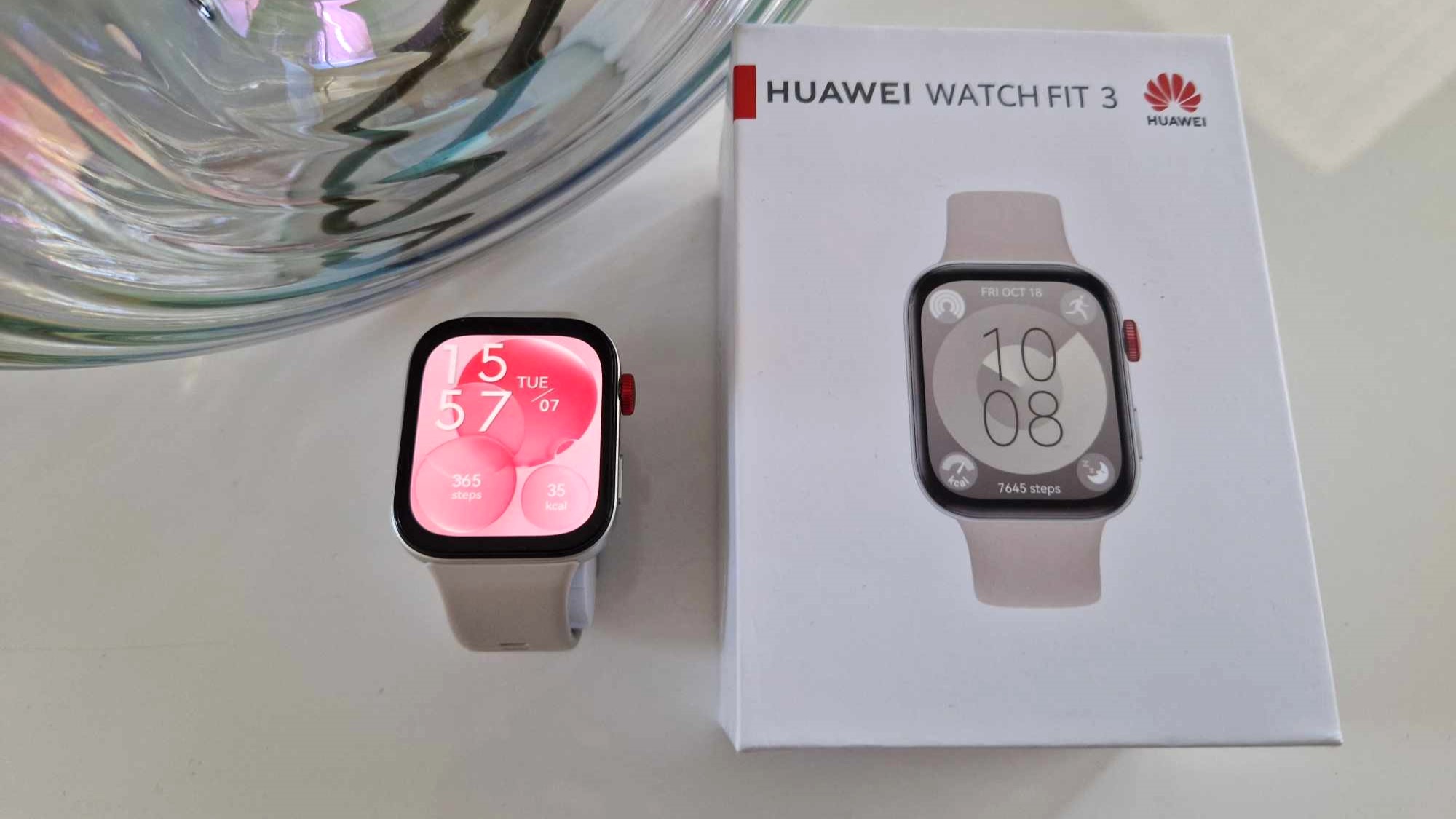 En hvit Huawei Watch Fit 3 ligger ved siden av den medfølgende emballasjen på et hvitt bord.