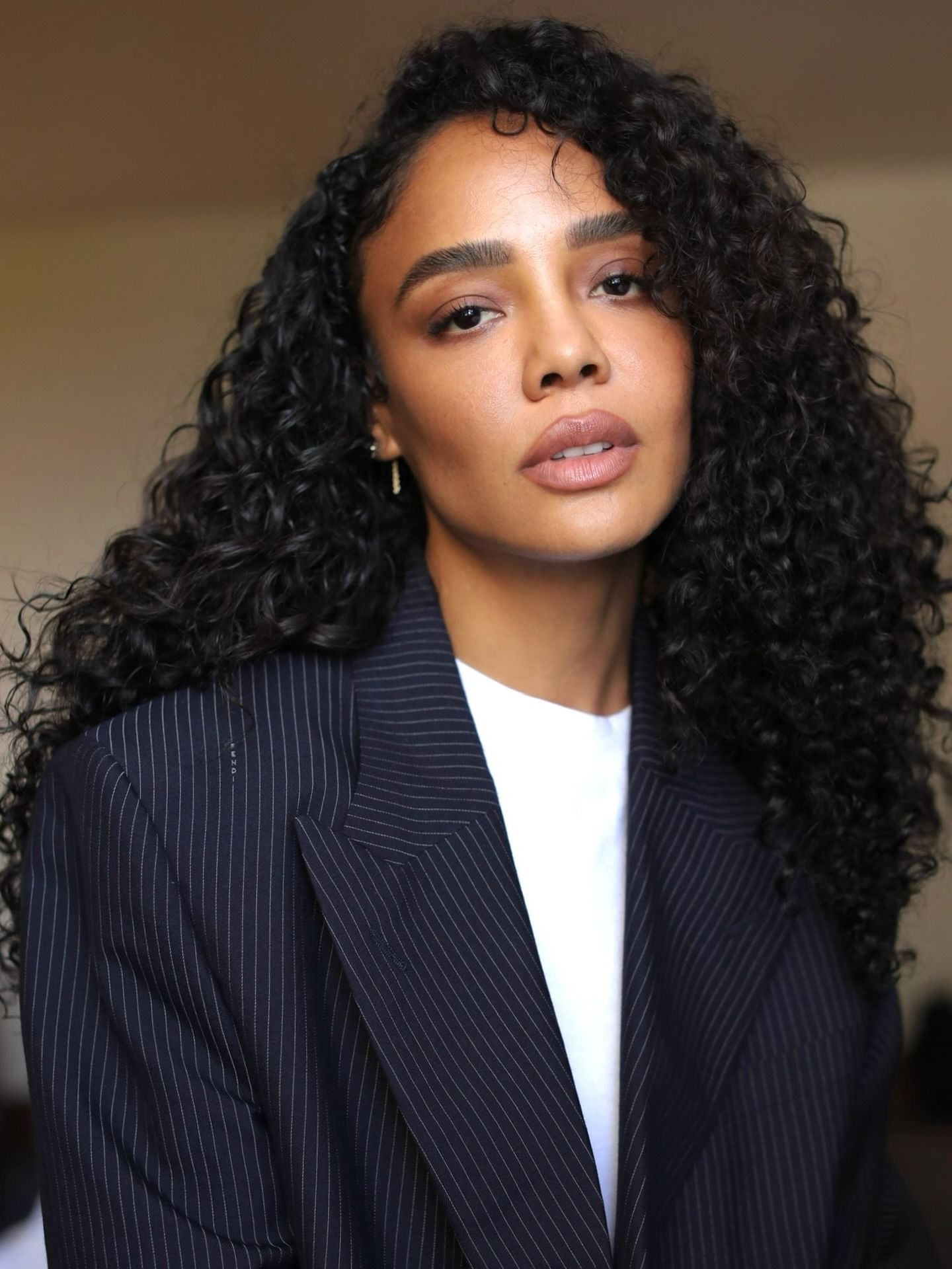 Tessa Thompson with dark brunette curls wearing a white T-shirt and navy striped blazer