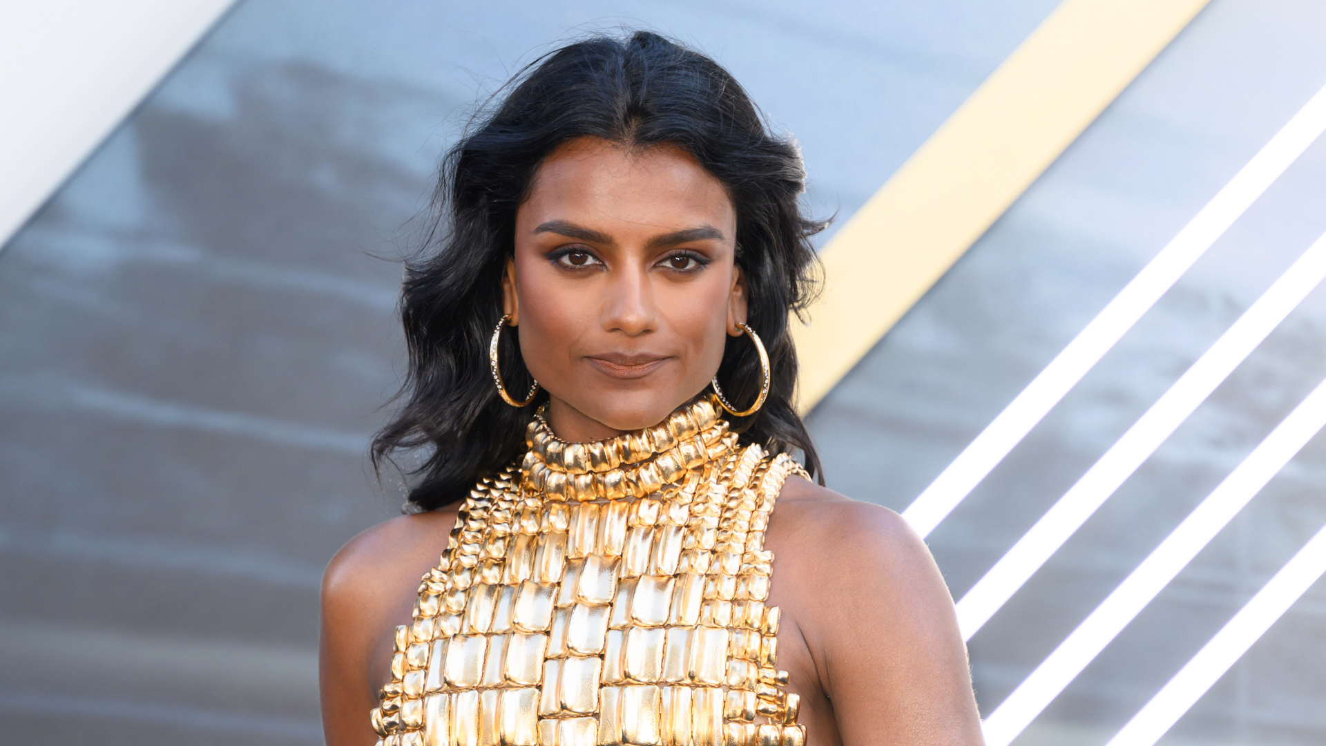 Simone Ashley wearing a gold halter neck top. Her hair is curled and she is also wearing gold hoop earrings. 