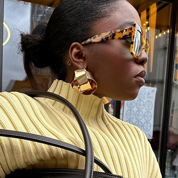 a woman wearing a butter yellow sweater, brown leather bag, tortoise shell sunglasses, and gold earrings