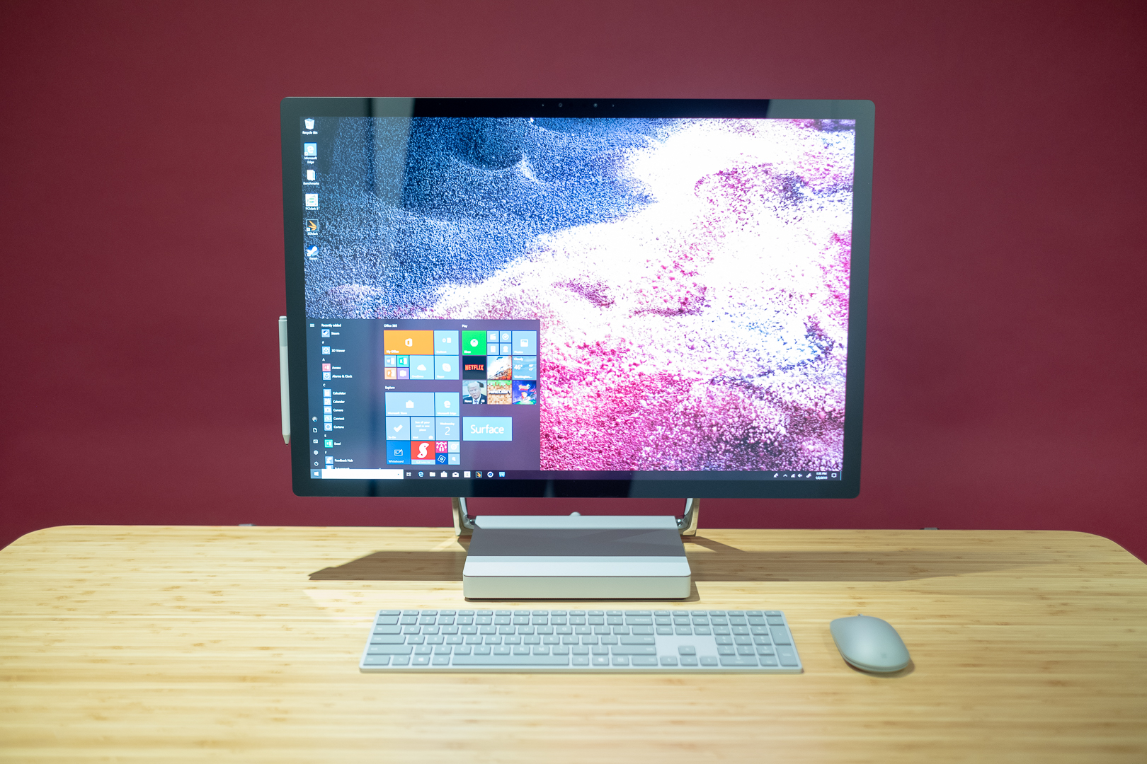 En Surface Studio 2 står på ett träfärgat skrivbord framför en vinröd vägg, med en datormus och ett tangentbord framför.