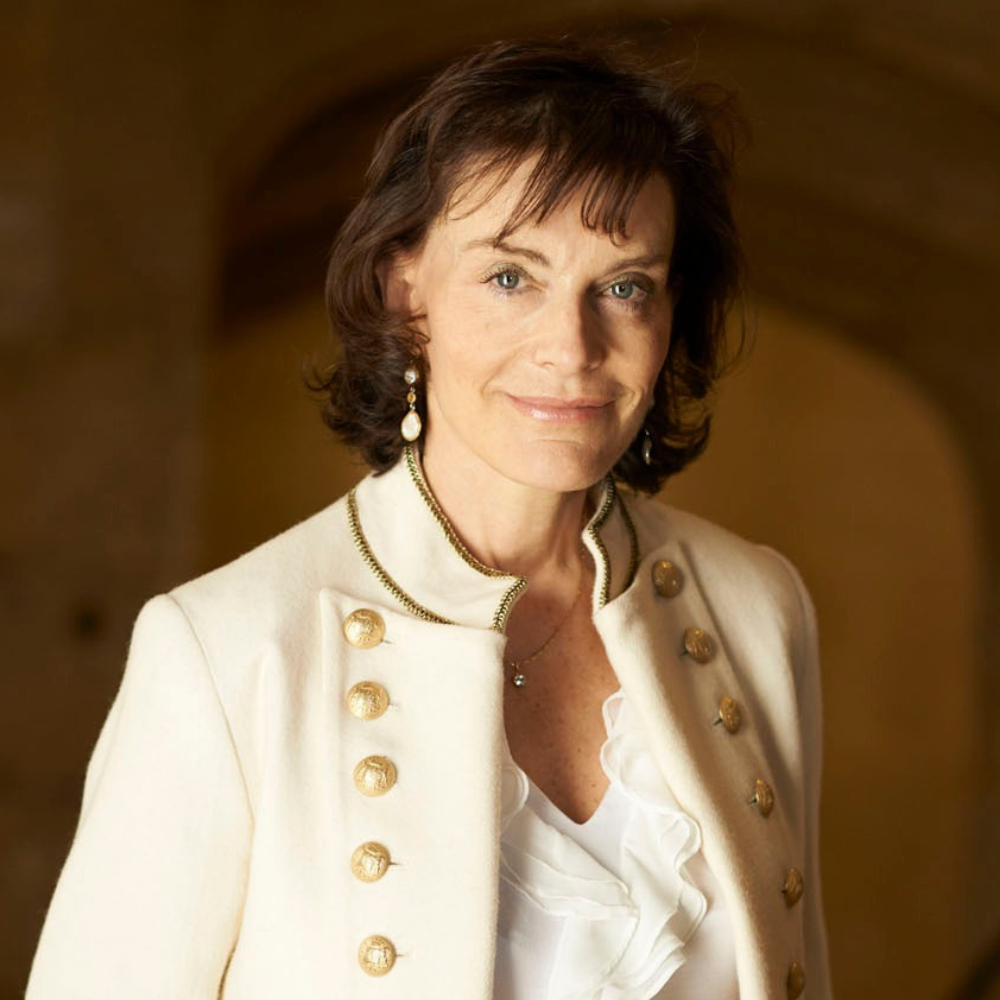 Woman with short dark hair wearing a cream military-style jacket with gold buttons, in a warmly lit archway.