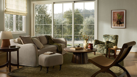 a living room with a taupe couch, a wooden coffee table, a brown sculptural chair, and a patterned chair. The room features a wall of windows and an artwork hanging from the wall