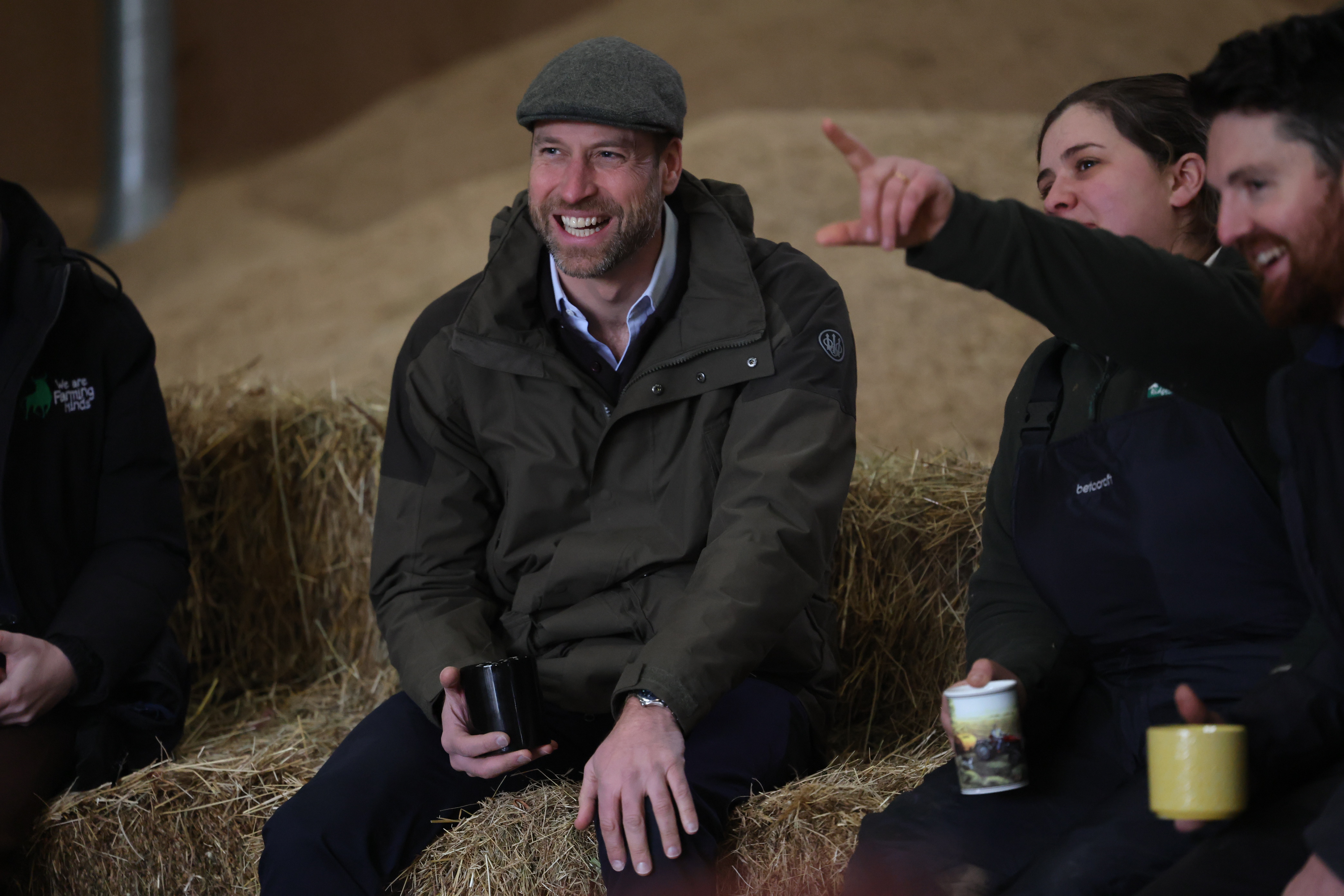 Prince William wearing a green cap and jacket sitting on a bale of hay with people drinking tea