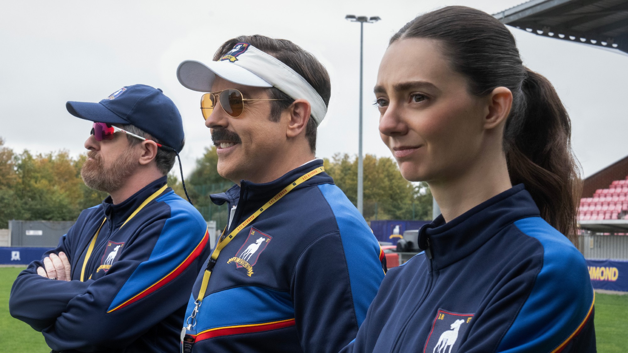 From left to right: Brendan Hunt, Jason Sudeikis and Tanya Reynolds standing together on the pitch.