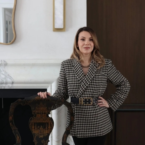 Image of a woman i a houndstooth blazer standing in front of a white marble fireplace with her hand resting on the top of an antique wooden chair.