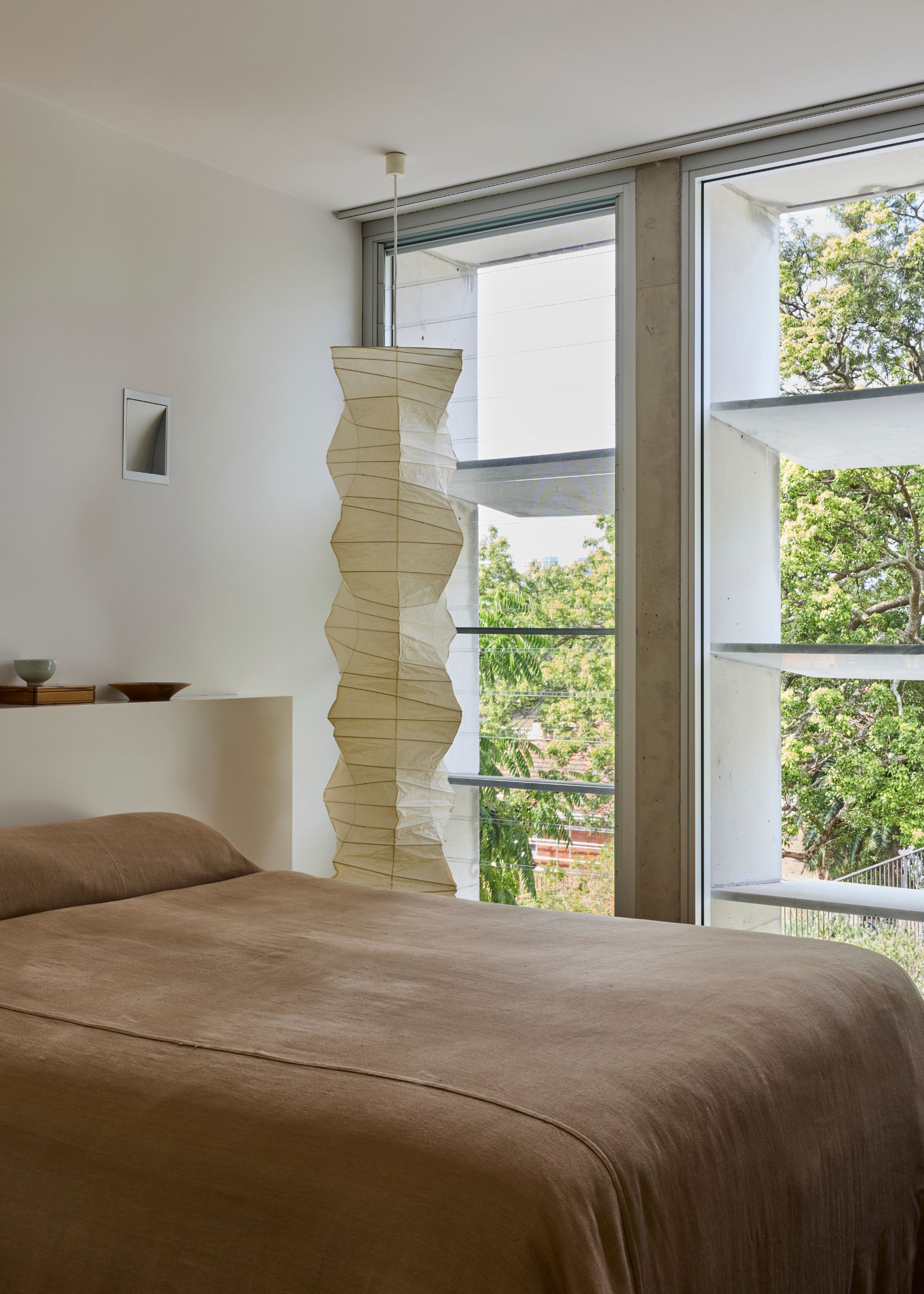 A white bedroom with large slatted windows, a pendant paper lamp, and brown bedding