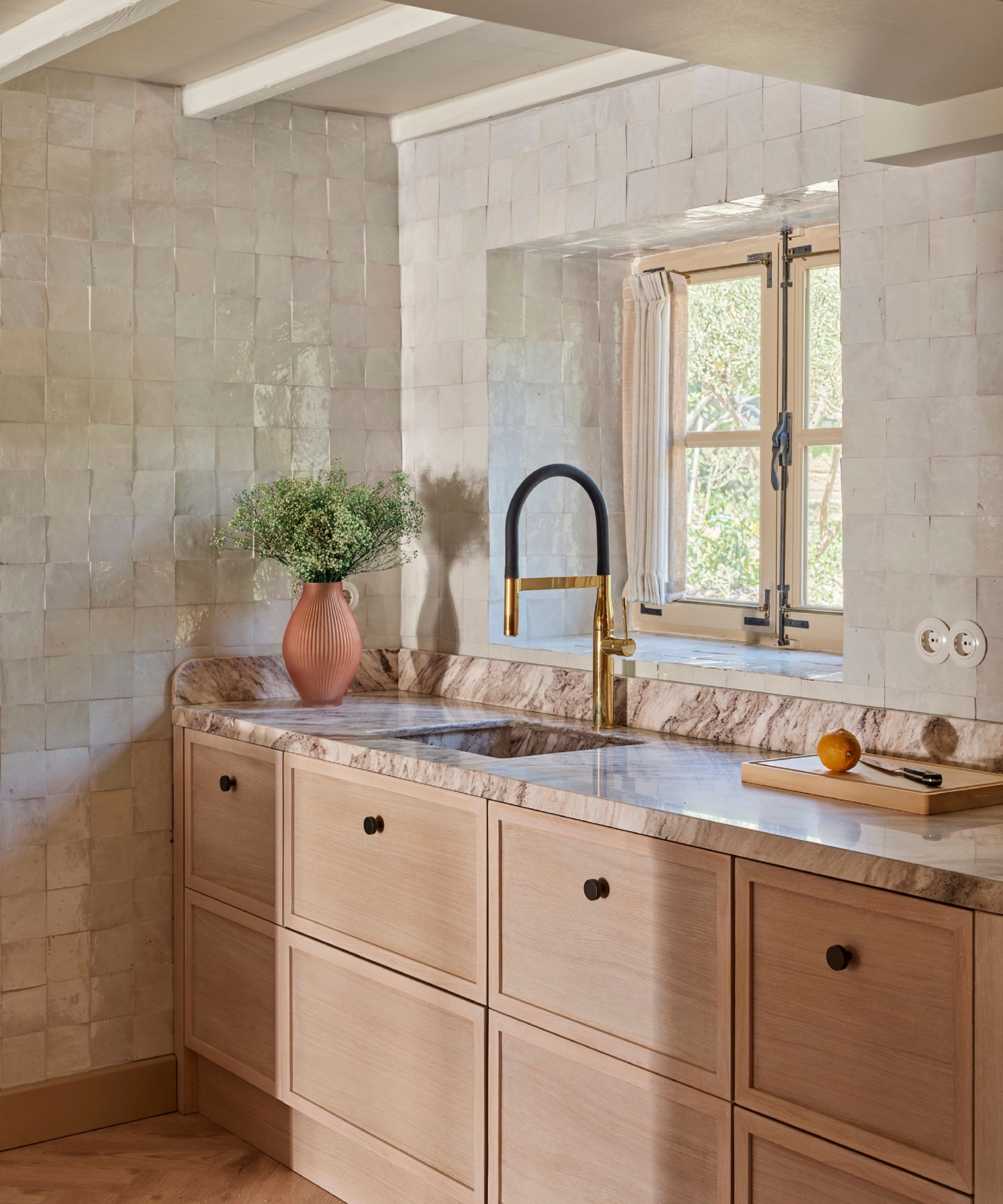 Warm, elegant kitchen with light wood cabinets, marble worktops and cream-toned tiled walls. A brass tap stands beneath a window, with natural light reflecting off textured tiles, creating a calm, refined atmosphere with natural materials.