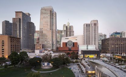 View of The San Francisco Museum of Modern Art (SFMoMA) building under a clear sky. There are multiple buildings surrounding the museum and Yerba Buena Gardens is nearby