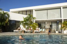 Man is swimming in a pool at a mansion and several friends tanning on the pool deck.