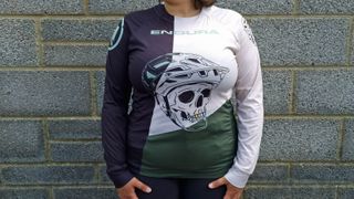 A long sleeve mountain biking jersey with a black panel on the left, a green panel on the lower right, and a white panel on the upper right, with a print of a skull wearing a helmet overlaid in black