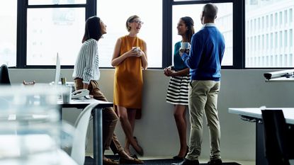 Four coworkers enjoy coffee together standing by an office window