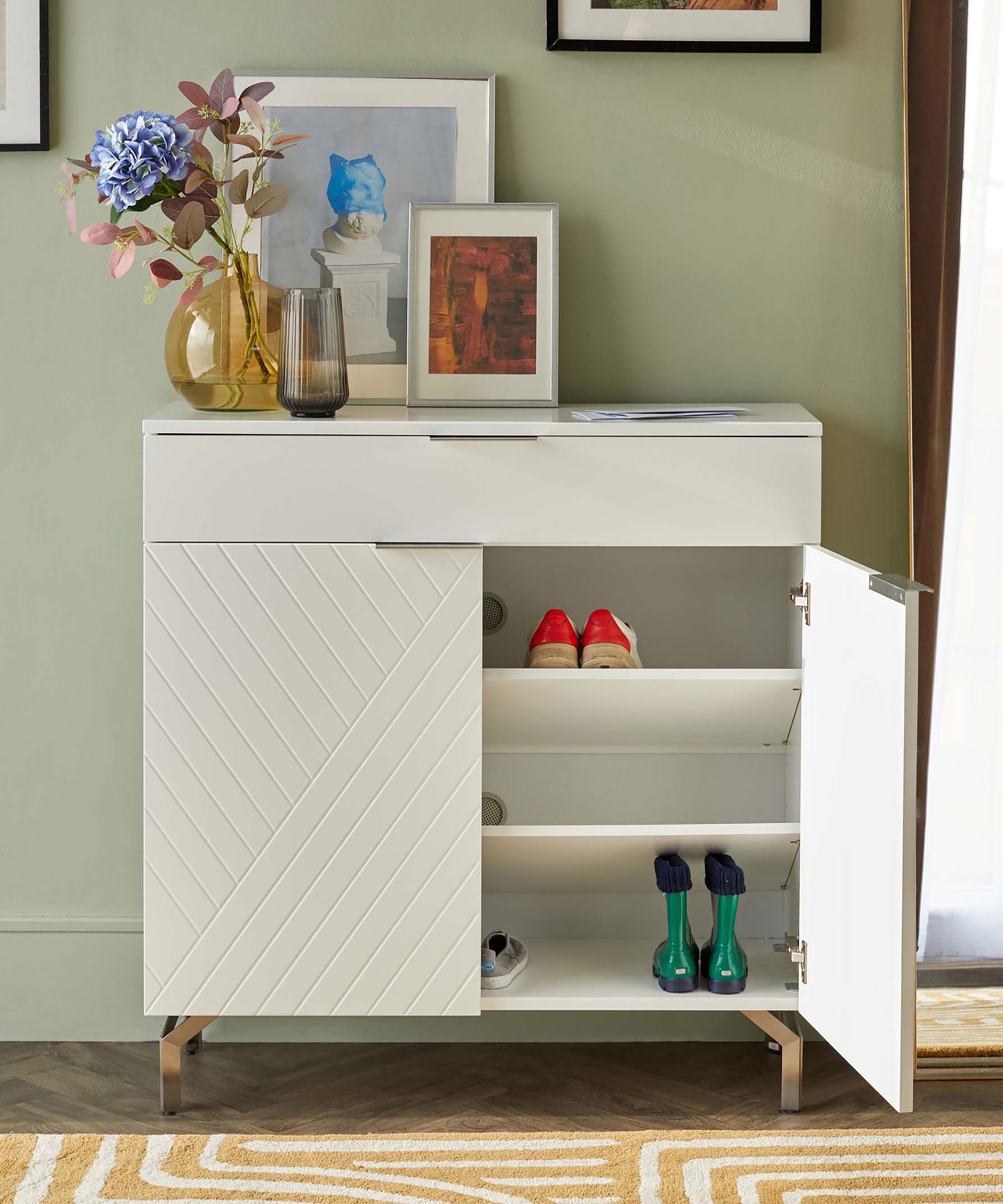 A white gloss shoe cabinet with chevron-patterned doors, one side open to reveal two internal shelves holding red heels and green wellington boots, topped with framed artwork, a flower vase and decorative objects against a sage green wall.