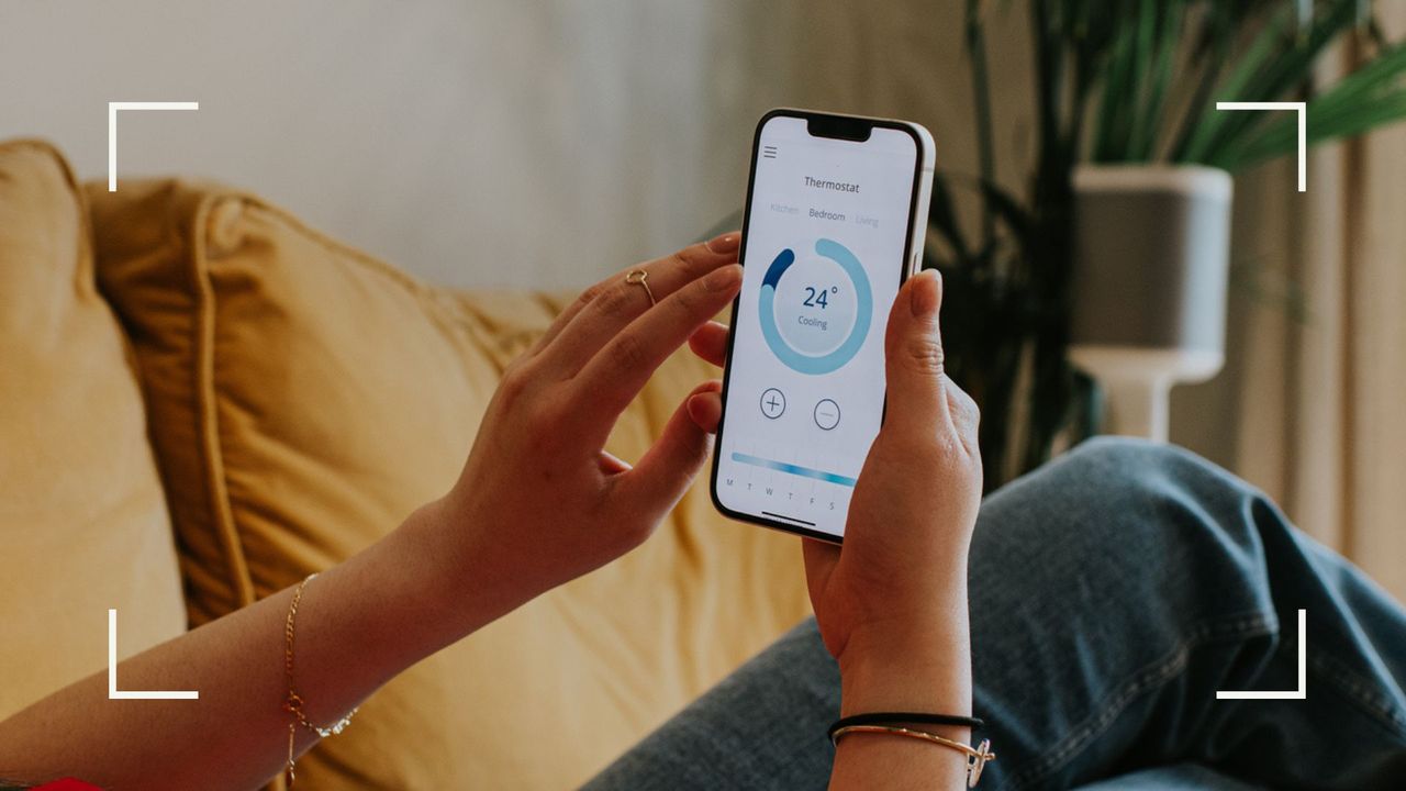 Woman lying down on yellow sofa in living room holding smartphone checking the thermostat on an app after considering whether to leave the heating on overnight