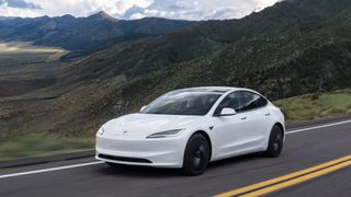 A white Tesla Model 3 driving along a winding mountain highway under a cloudy sky.