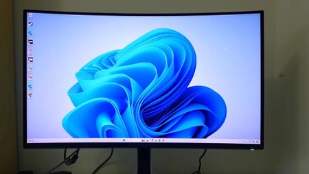 A desktop monitor displays the blue abstract Windows 11 wallpaper, with icons along the left edge; the clean, bright screen contrasts with a dim room.