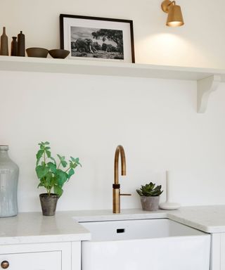 A white kitchen with a long wall shelf with a black and white print and golden wall sconce, with a sink below it with a gold curved tap, a potted basil plant and succulent next to it