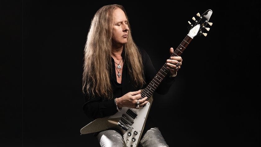 Jerry Cantrell poses with his Gibson Flying V. He wears a long black t-shirt and silver pants.