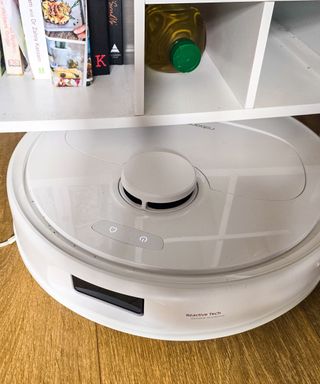 A white robot vacuum underneath a white bar cart with books and bottles on it, on top of a brown wooden floor