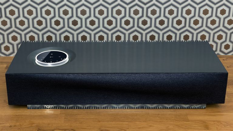 Best wireless speakers, image shows Naim Mu-so 2 sitting on wooden bench with patterned wallpaper behind