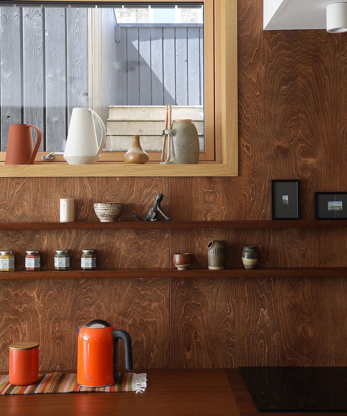 A kitchen with dark wooden walls and surfaces. There are small, shallow built-in shelves containing trinkets and small clay pots. The orange kettle and biscuit tin give a pop of colour and are a nod to the mid century modern design of the house