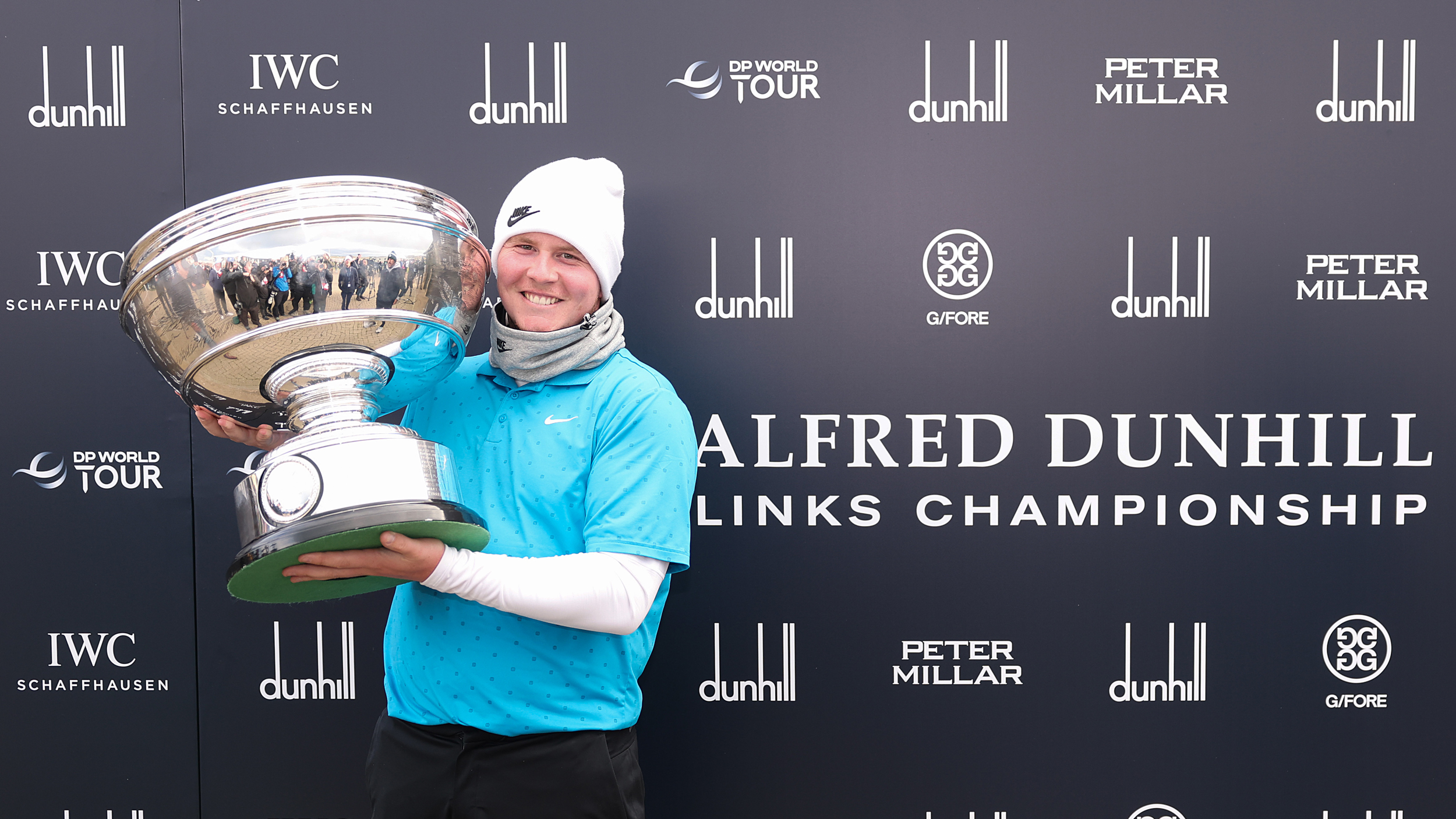 Robert MacIntyre poses with the Alfred Dunhill Links Championship trophy beside a large sponsor-filled sign with the event name on