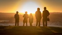 five silhouetted figures stand on a hill as the glare of the rising sun fills the sky, illuminating the misty landscape below.