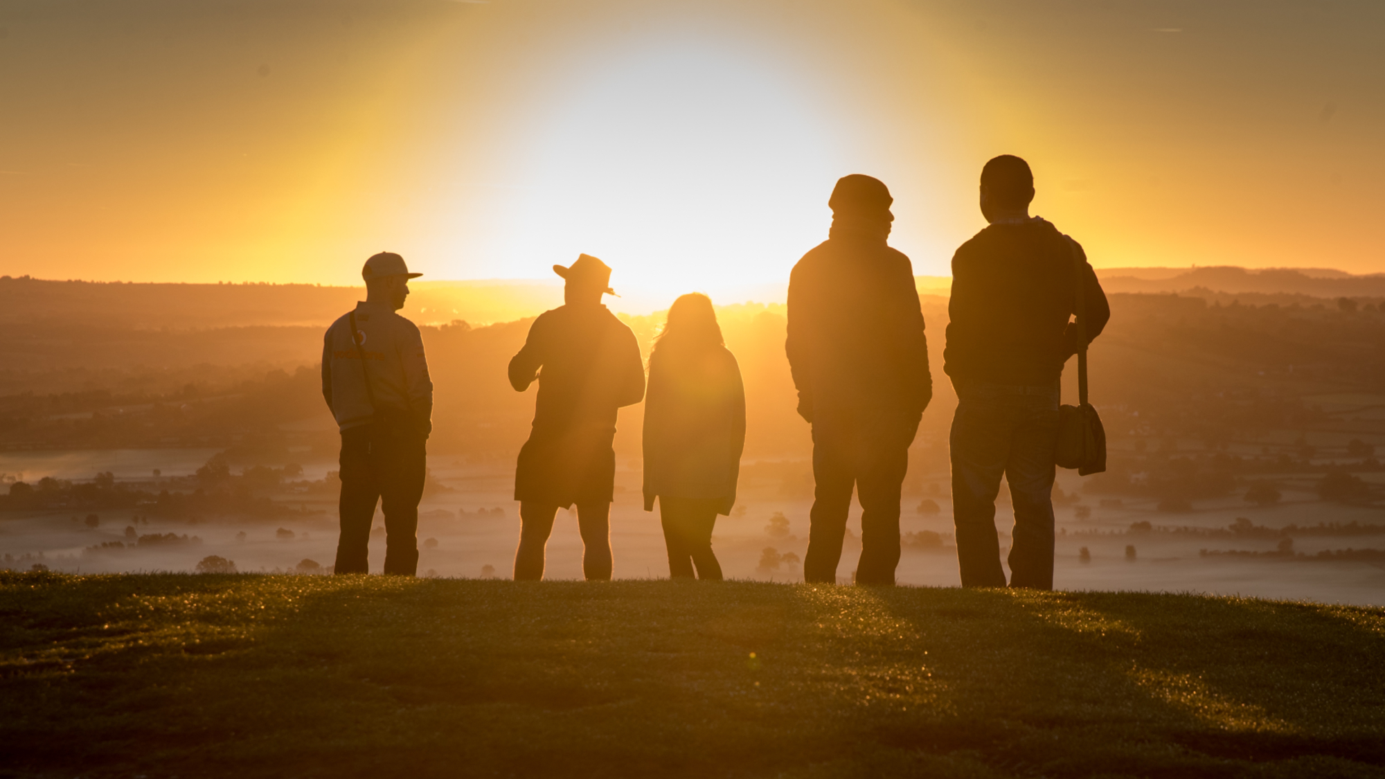 five silhouetted figures stand on a hill as the glare of the rising sun fills the sky, illuminating the misty landscape below.