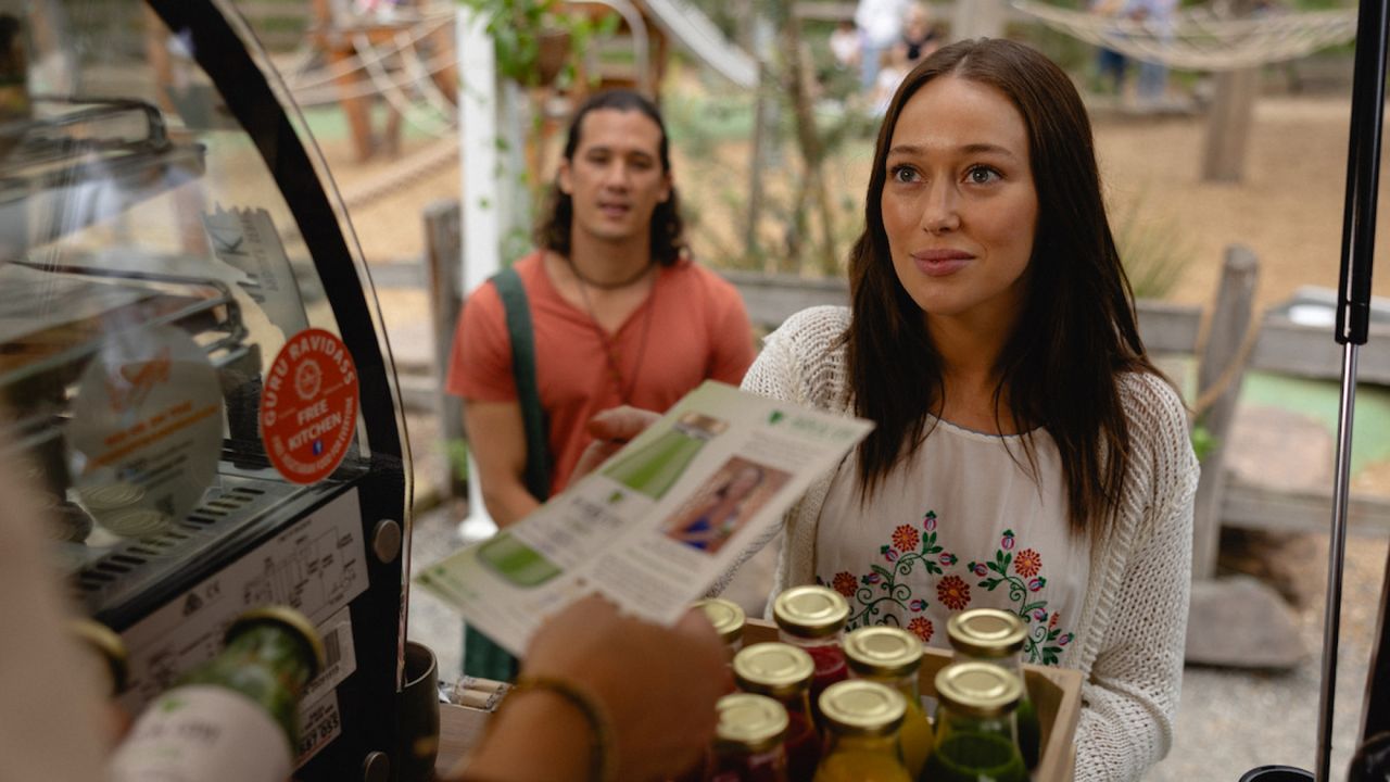 A woman (Alycia Debnam-Carey as Milla Blake) holds a basket of juice bottles as a man (Chai Hansen as Arlo) stands behind her. To the left, a hand holds a pamphlet about the juices. A still from &#039;Apple Cider Vinegar.&#039;