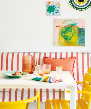 Bright and colorful dining area with striped, upholstered bench seat, orange cushions, white dining table, yellow chairs, artwork on walls