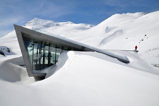 Close up view of the snow covered Trollstigen Visitor Centre under a blue sky. There is a person in a red jacket standing on the building and the mountains are also covered in snow