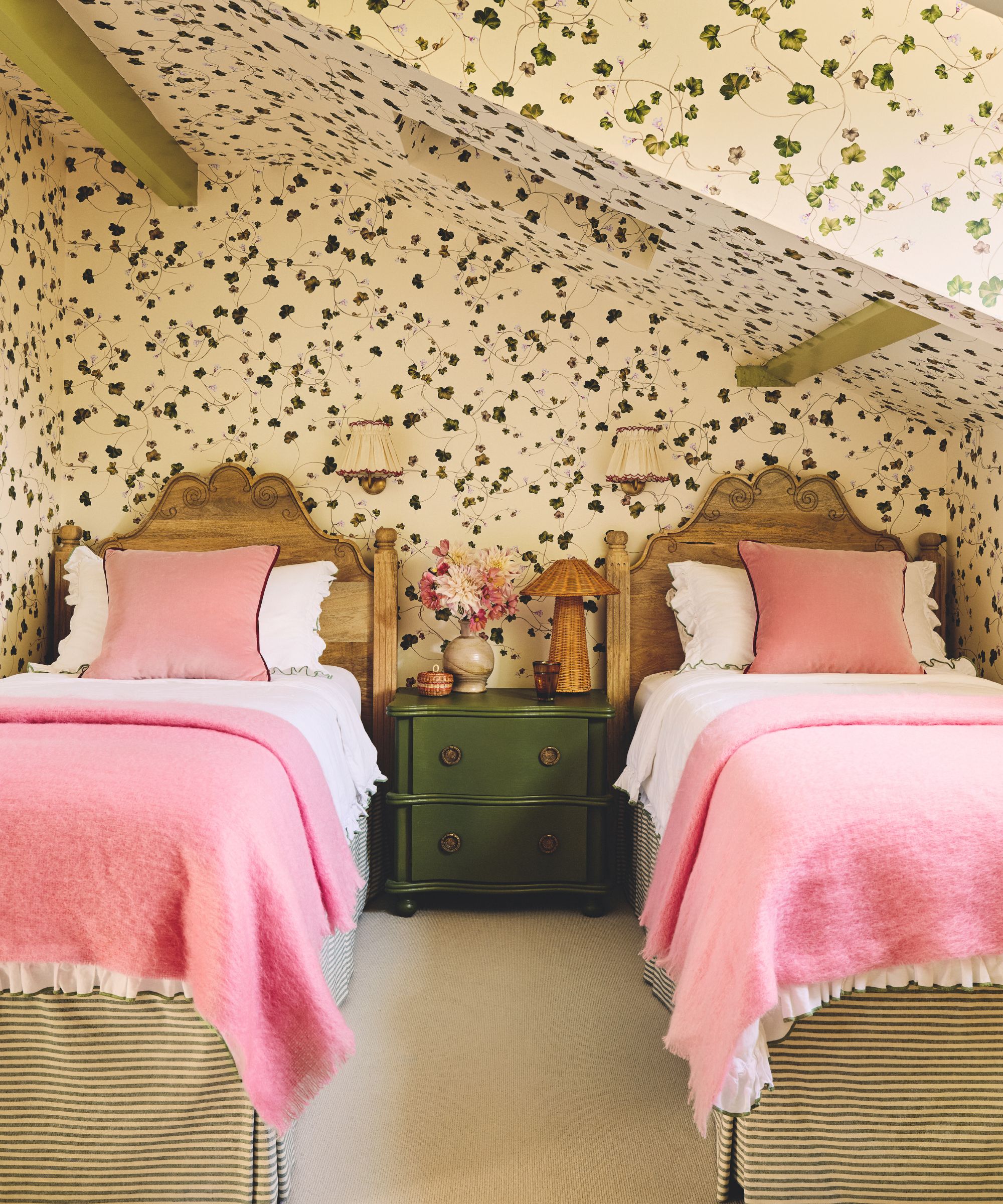 attic twin bedroom witch trailing ivy printed wallpaper on all walls and ceiling with green painted ceiling beams and a green nightstand and pink bed linens