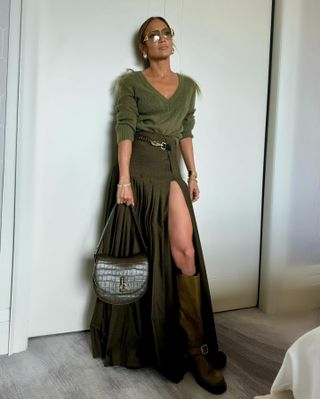 Jennifer Lopez wears a green v-neck sweater, green maxi pleated skirt, brown knee-high moto boots, a Burberry croc-effect handbag, gold earrings, and aviator sunglasses.