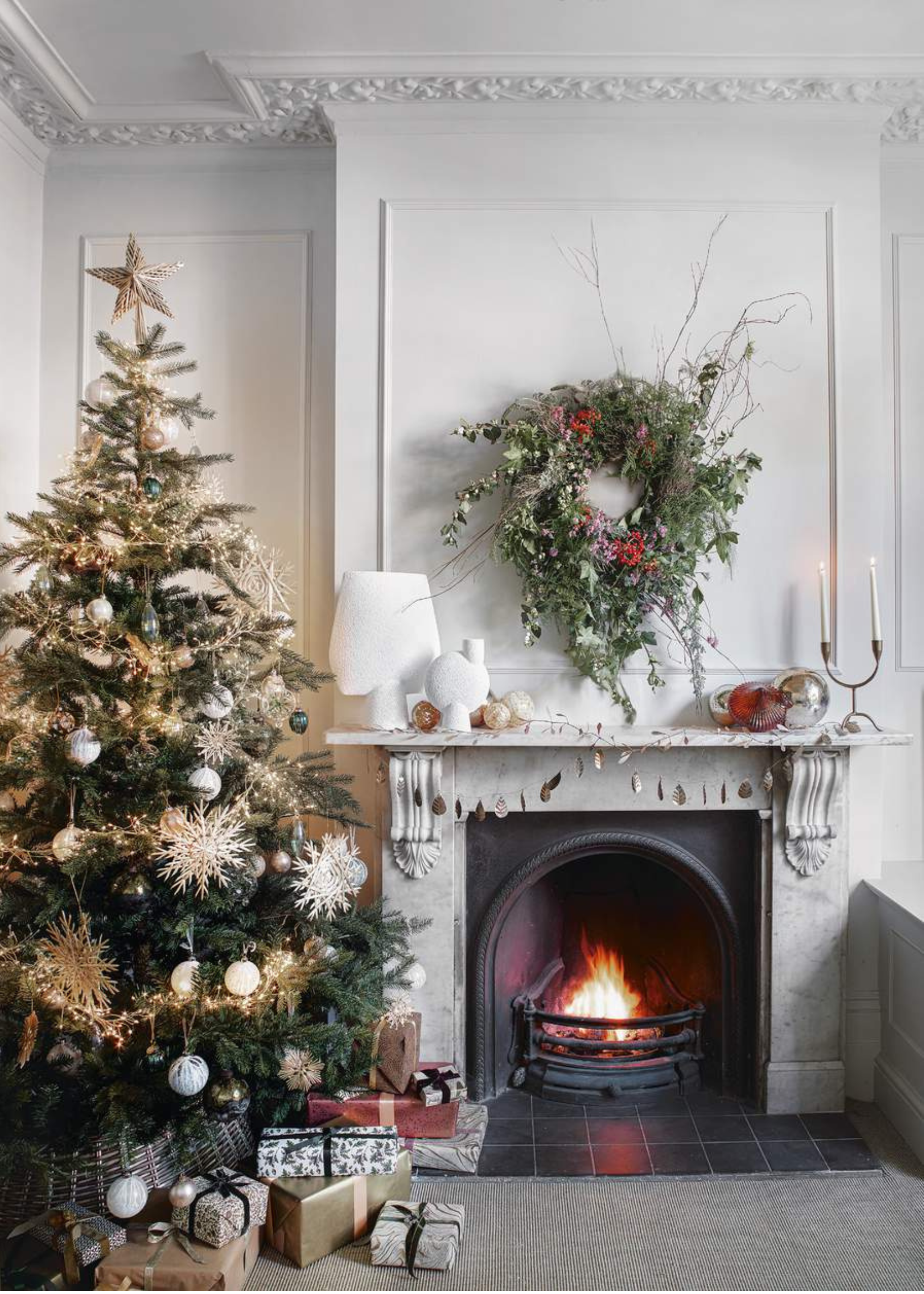 A white-walled living room with a large decorated christmas tree sitting next to a fireplace has a foraged floral wreath hanging like a piece of art above a lit fire