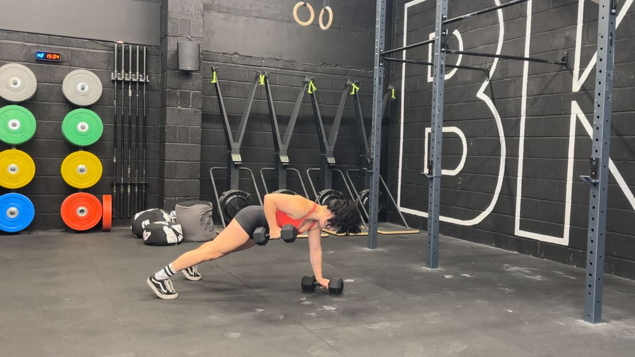 Raquel Sanjurjo performs a renegade row into triceps kickback in a gym. She is in a high plank position holding a pair of dumbbells. One is on the floor, the other has been rowed up to her right hip with her bent right elbow.