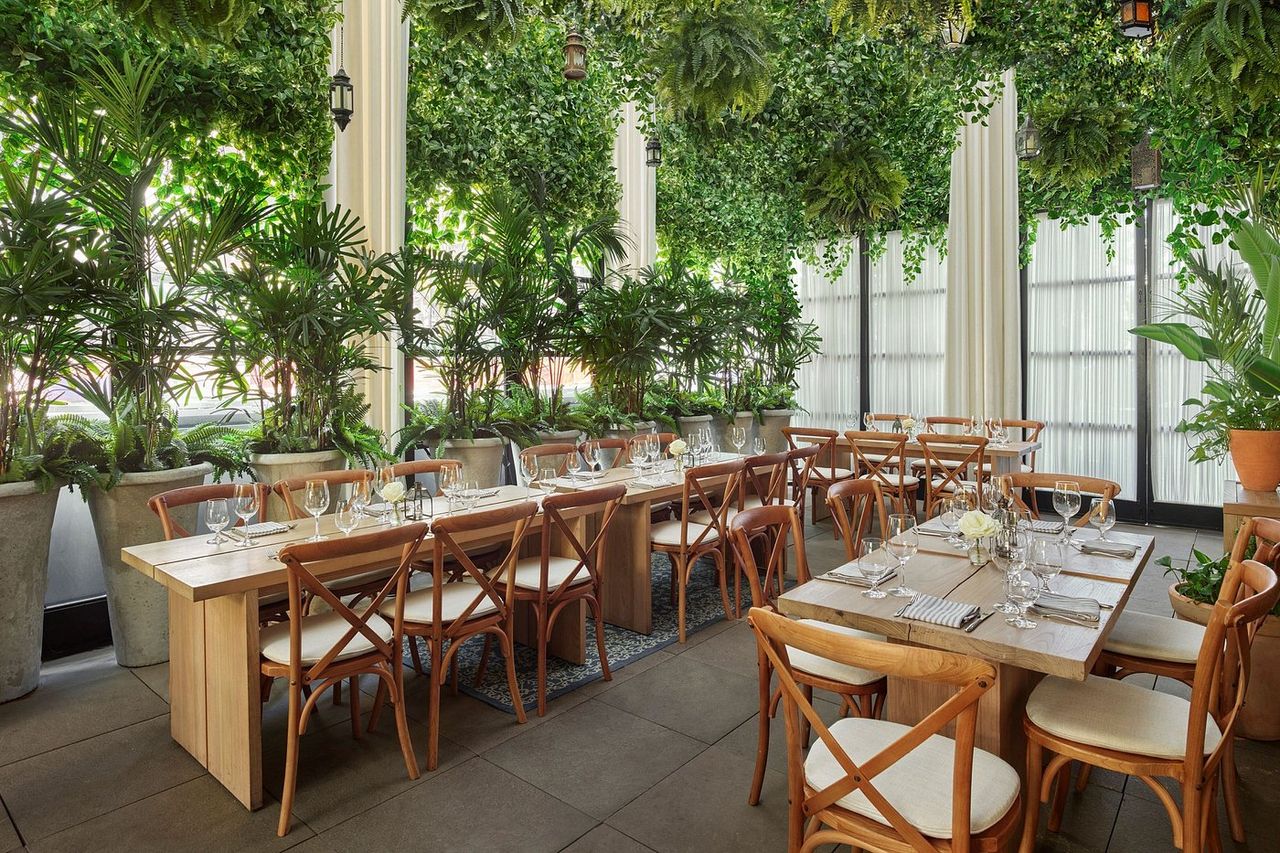A superbly decorated, hyper-modern hotel features minimalist cozy furniture including neutrally-hued large couches, standing lamps, tiled floors, cream curtains, wall-to-wall windows, and breathtaking views of the city.