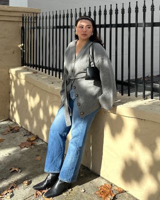 Fashion influencer Marina Torres poses on a sidewalk wearing a headband, chunky two-tone earrings, Freya black shoulder bag, gray belted cardigan sweater, straight-leg jeans, and black boots.