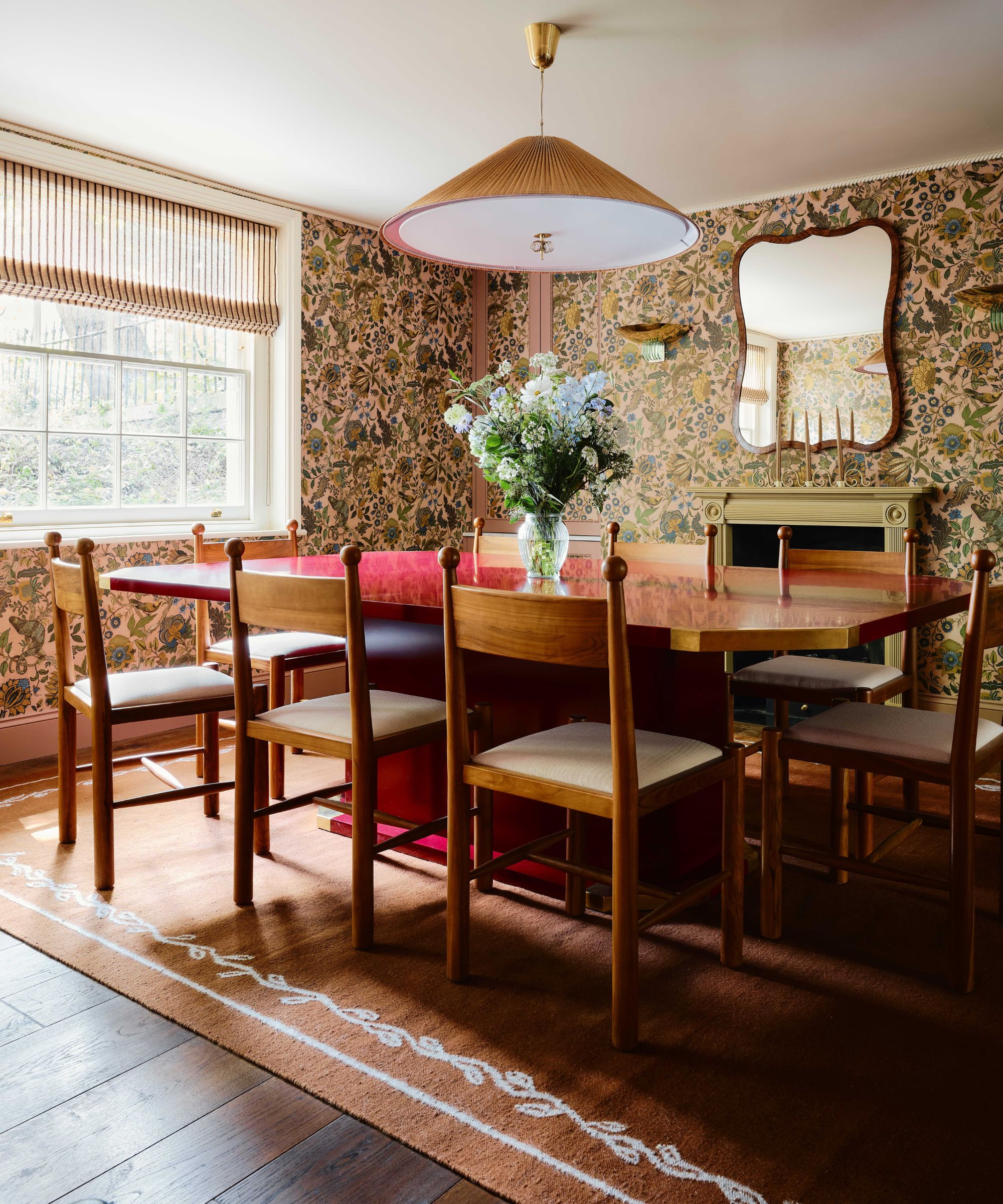large dining room with beige, green, blue floral wallpaper, dark wooden floor, a large rug and a red lacquered dining table with pine retro chairs