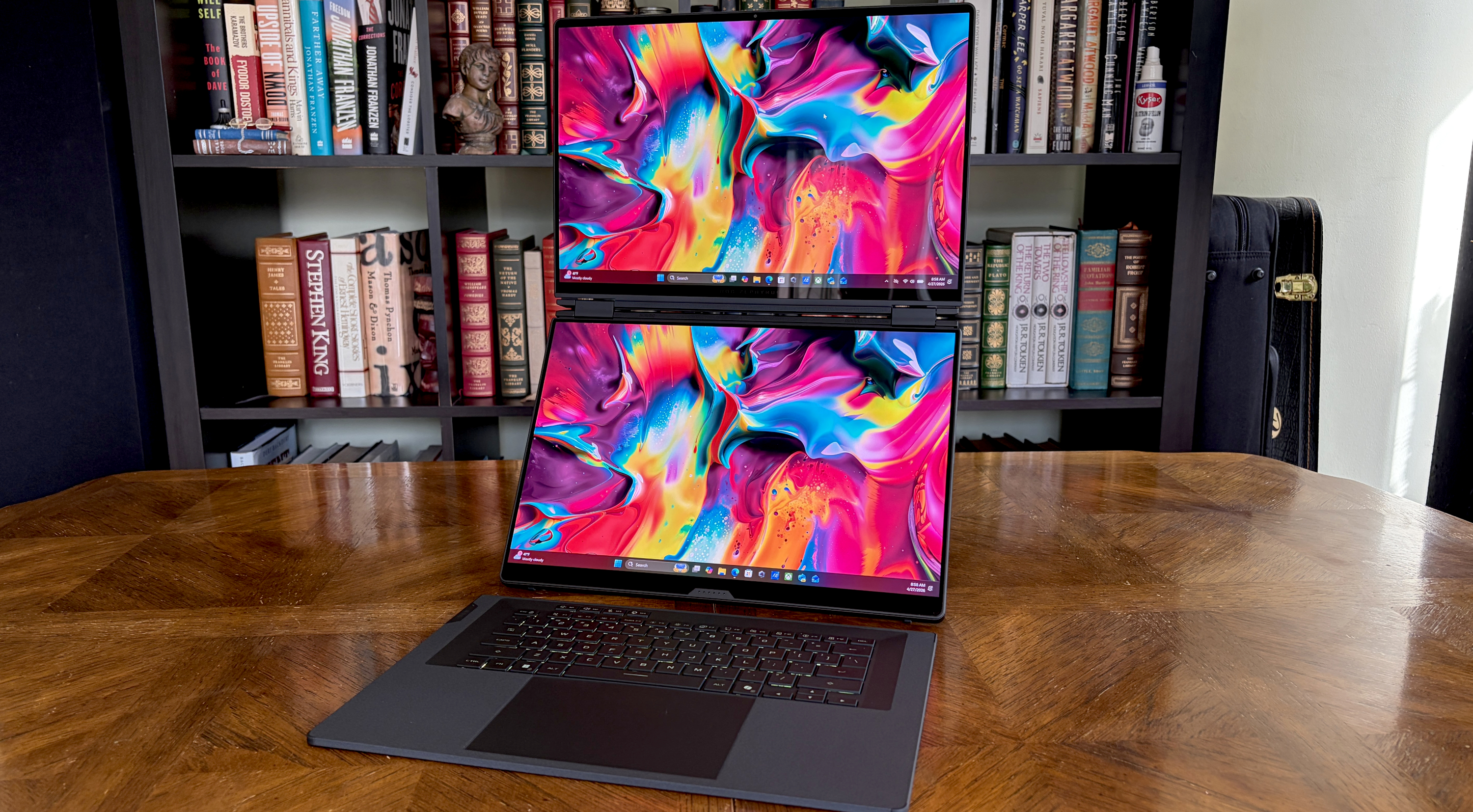 A laptop with a dual-screen setup on a wooden desk, displaying vibrant, abstract designs. Background features bookshelves filled with books, creating a focused and creative atmosphere.