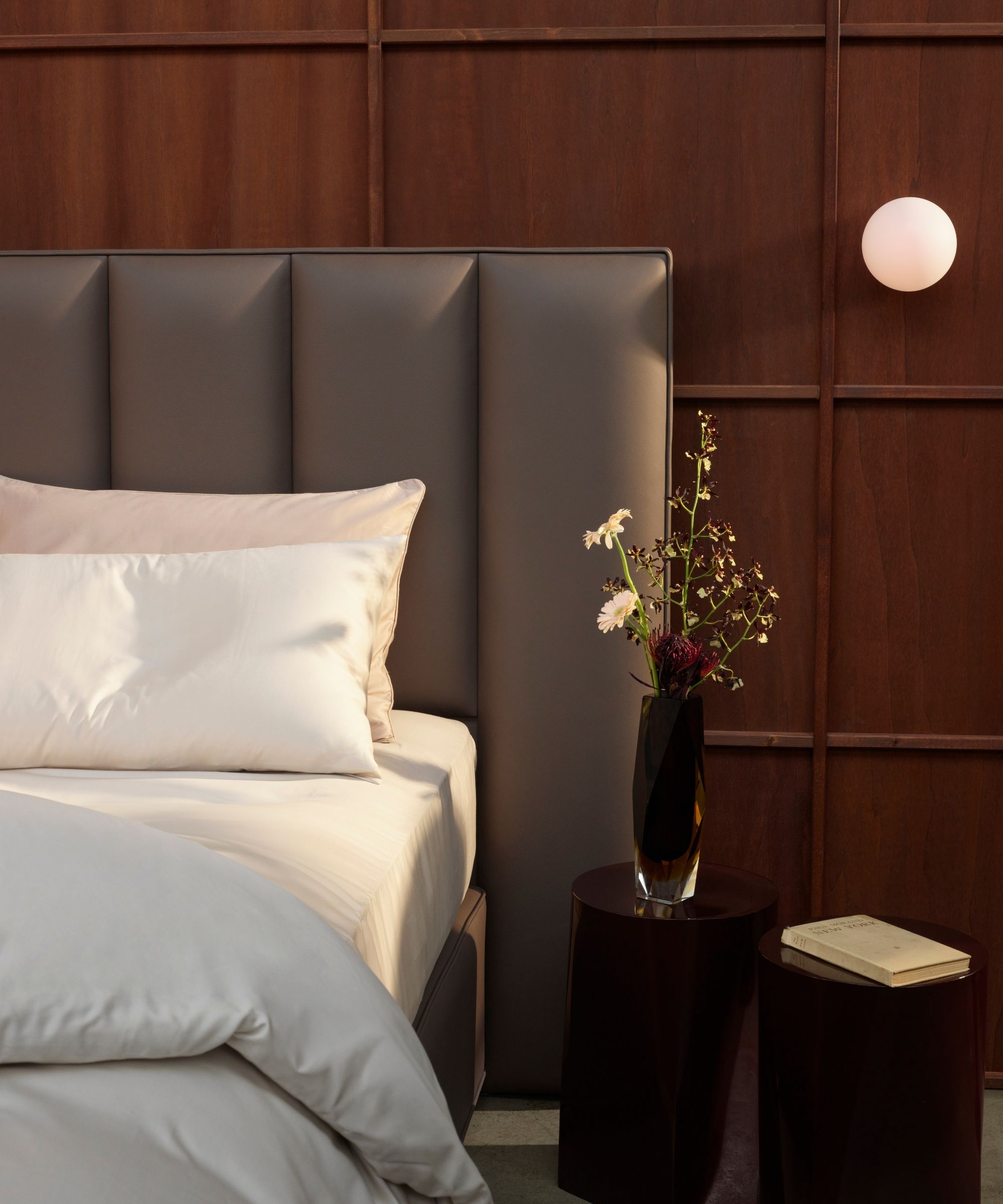 Dark wood panelled bedroom with a grey padded headboard, white bedding, dark vase with flowers, and a white book on two black circular nightstands, and a circular wall lamp.